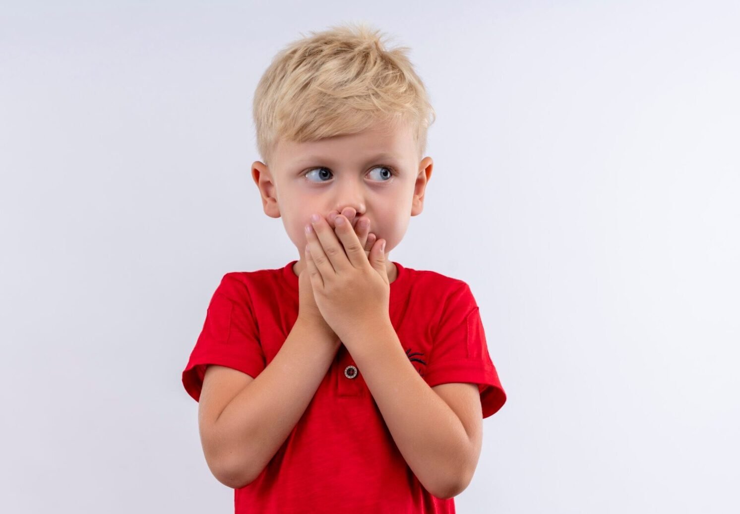 a surprised little cute blonde boy in red t-shirt keeping hands on his mouth while looking side on a white background