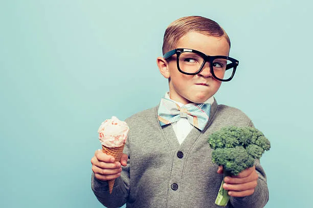 A young nerd boy with glasses is not sure whether to eat the ice cream cone or the broccoli. He is not sure which one is healthier.