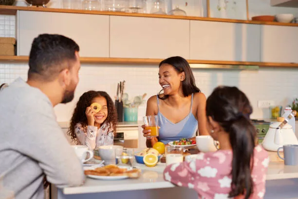 Cute little girl makes a funny face while looking with one eye through a biscuit with a hole during breakfast. Hispanic father and mother share a happy breakfast with their mixed race children in kitchen. Multiethnic family enjoying playful moments during their morning meal at home.