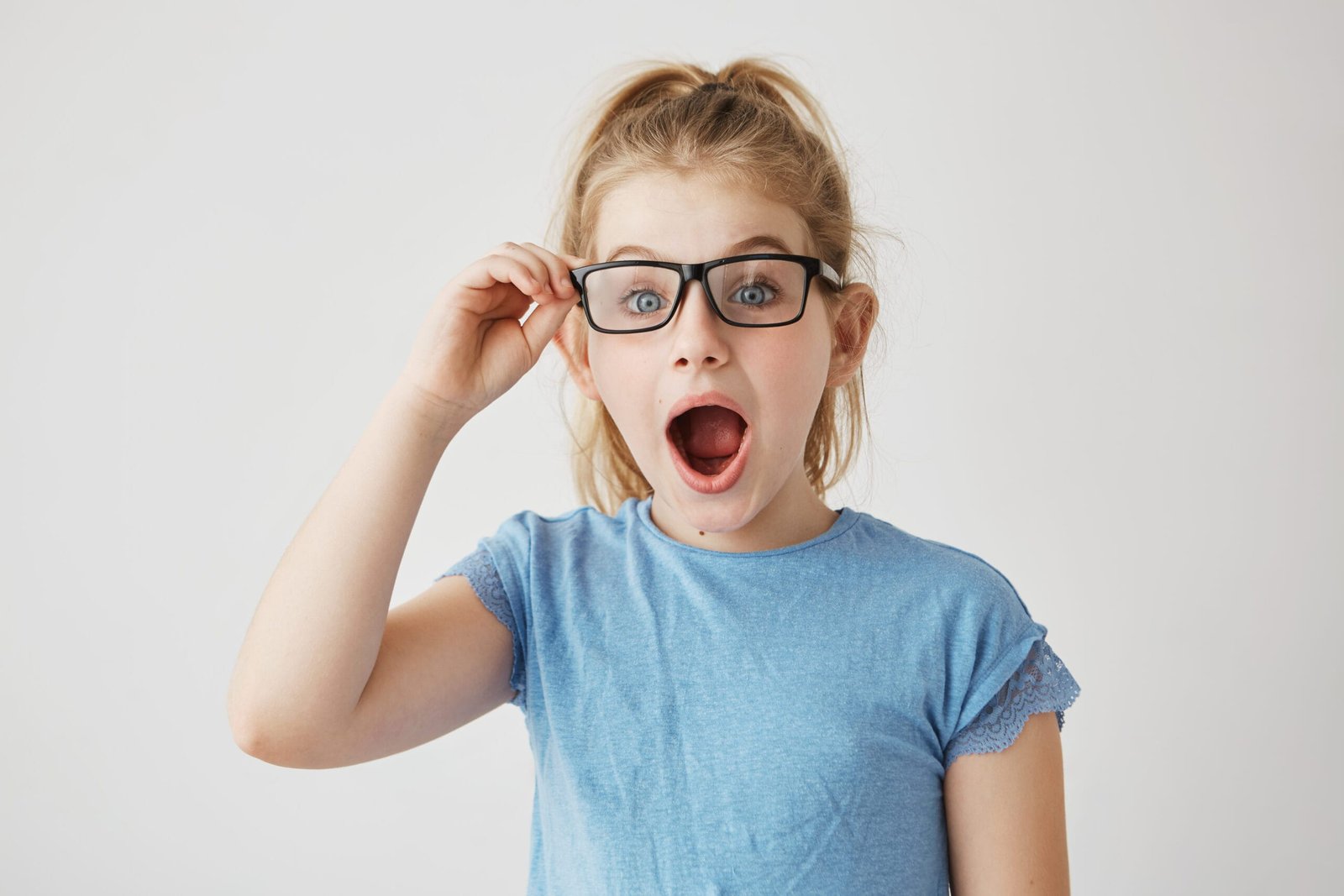 Little beautiful girl with bright blue eyes and blonde hair posing with opened mouth, holding her glasses with hand looking super surprised