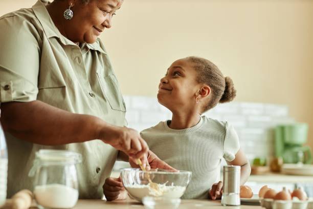 Side view of charming Black girl looking at loving grandma with adoring smile during cooking pancakes together at beige kitchen, copy space
