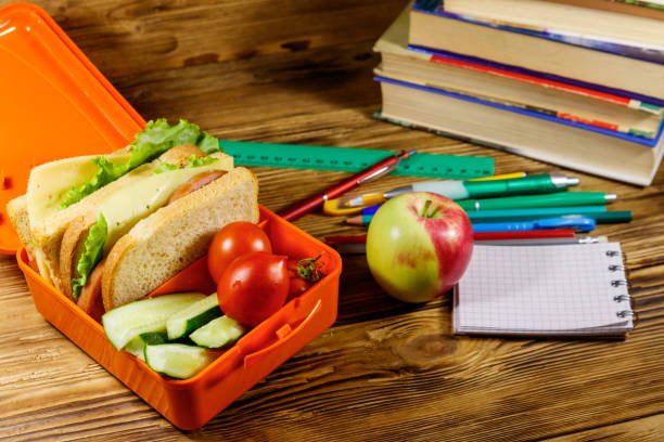 Back to school concept. School supplies, books, apple and lunch box with sandwiches and fresh vegetables on a wooden desk