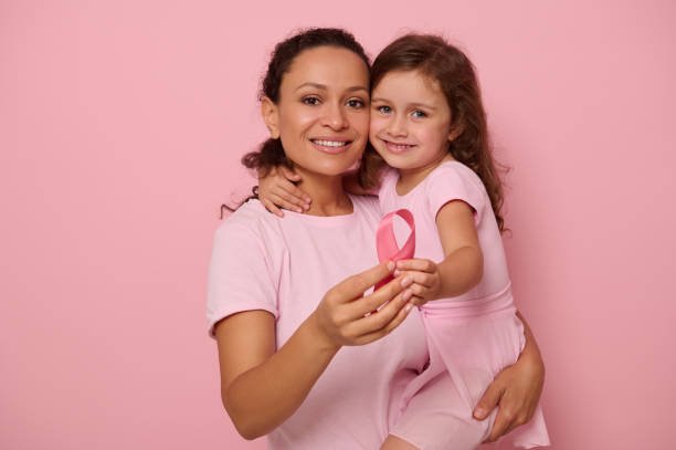 Two generations of women, woman and girl, mother and daughter posing against a pink background, holding a pink ribbon, showing support and solidarity to patients and cancer survivors. Medical awareness